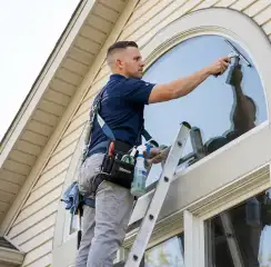 Professional window cleaner on a ladder using a squeegee to clean an exterior arched window, Raleigh, NC.